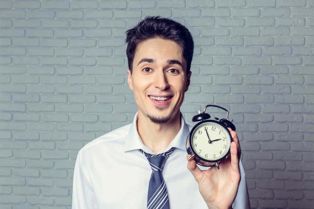 Uśmiechnięty mężczyzna pokazujący stary zegarek z budzikiem na tle ceglanej ściany